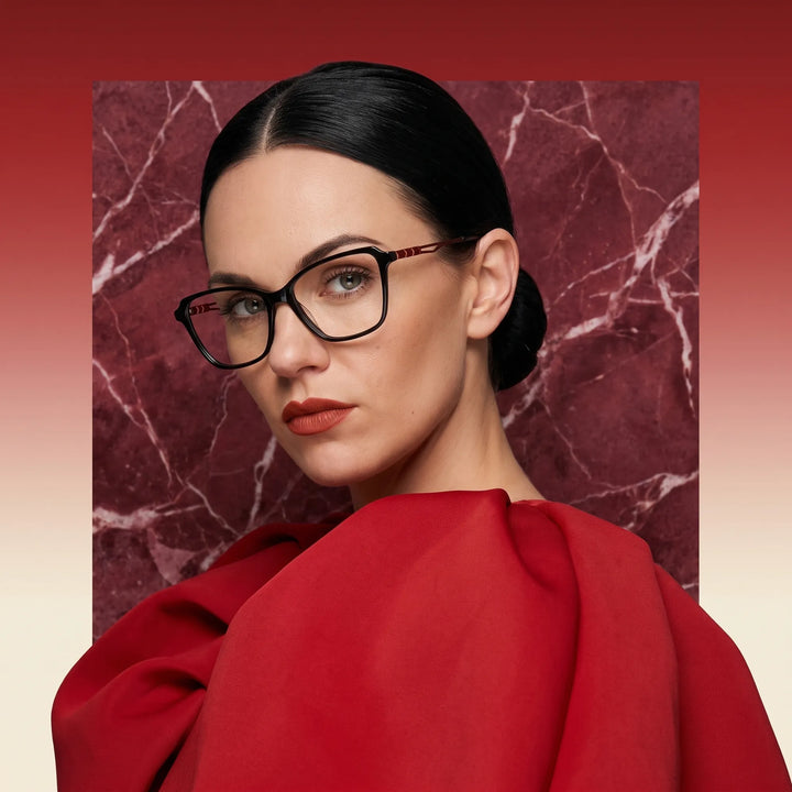 Anteojos Fiore 1006 en negro, en modelo – retrato de campaña, fondo de mármol rojo y vestido escarlata drapeado