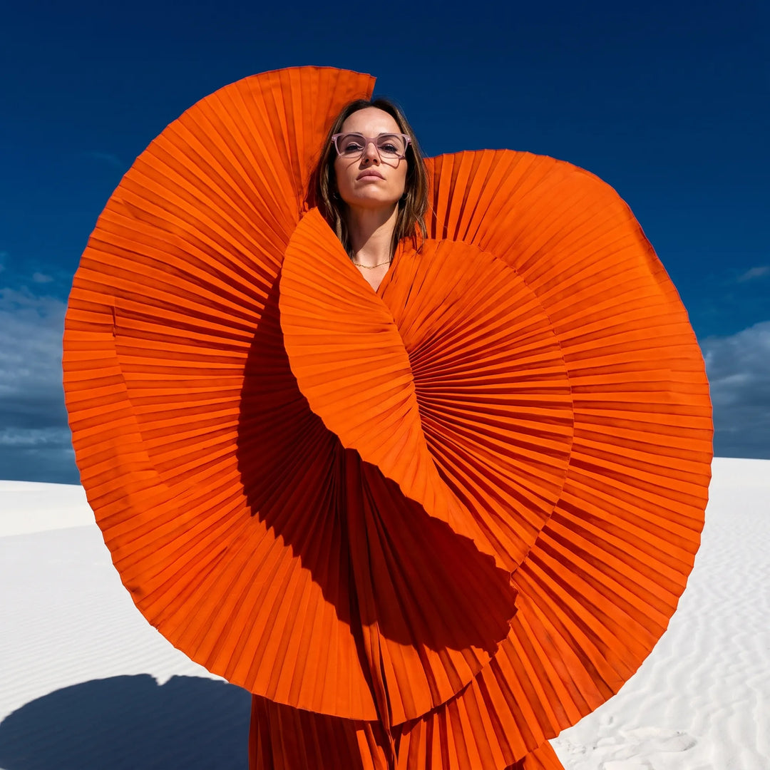 Anteojos Fiore 1006 en rosa, en modelo – retrato de campaña, abanico plisado naranja escultórico sobre dunas blancas