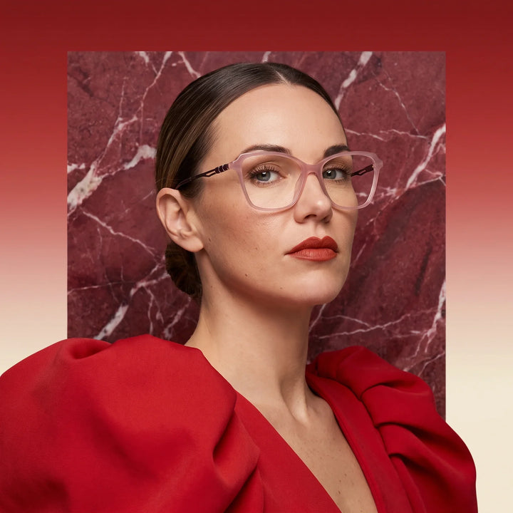 Anteojos Fiore 1006 en rosa, en modelo – retrato de campaña, fondo de mármol rojo y vestido escarlata drapeado