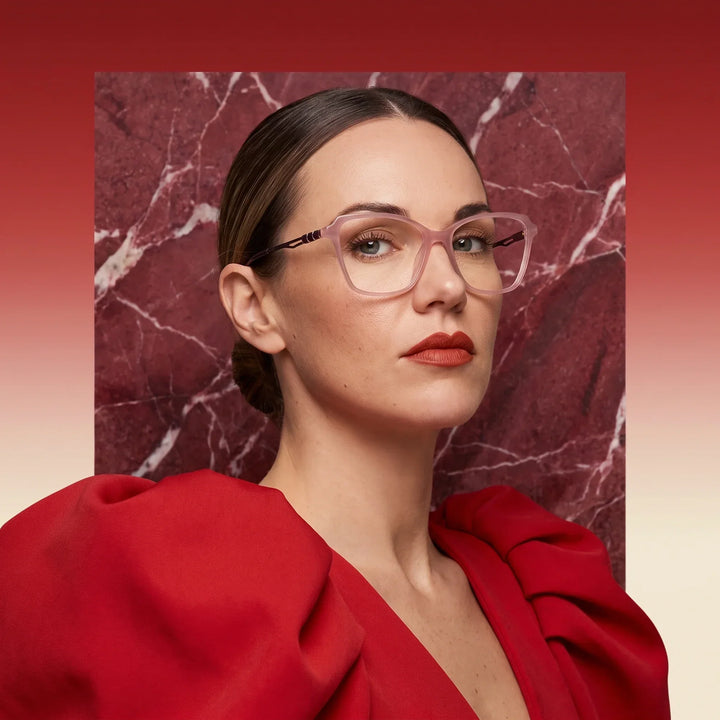 Anteojos Fiore 1006 en rosa, en modelo – retrato de campaña, fondo de mármol rojo y vestido escarlata drapeado