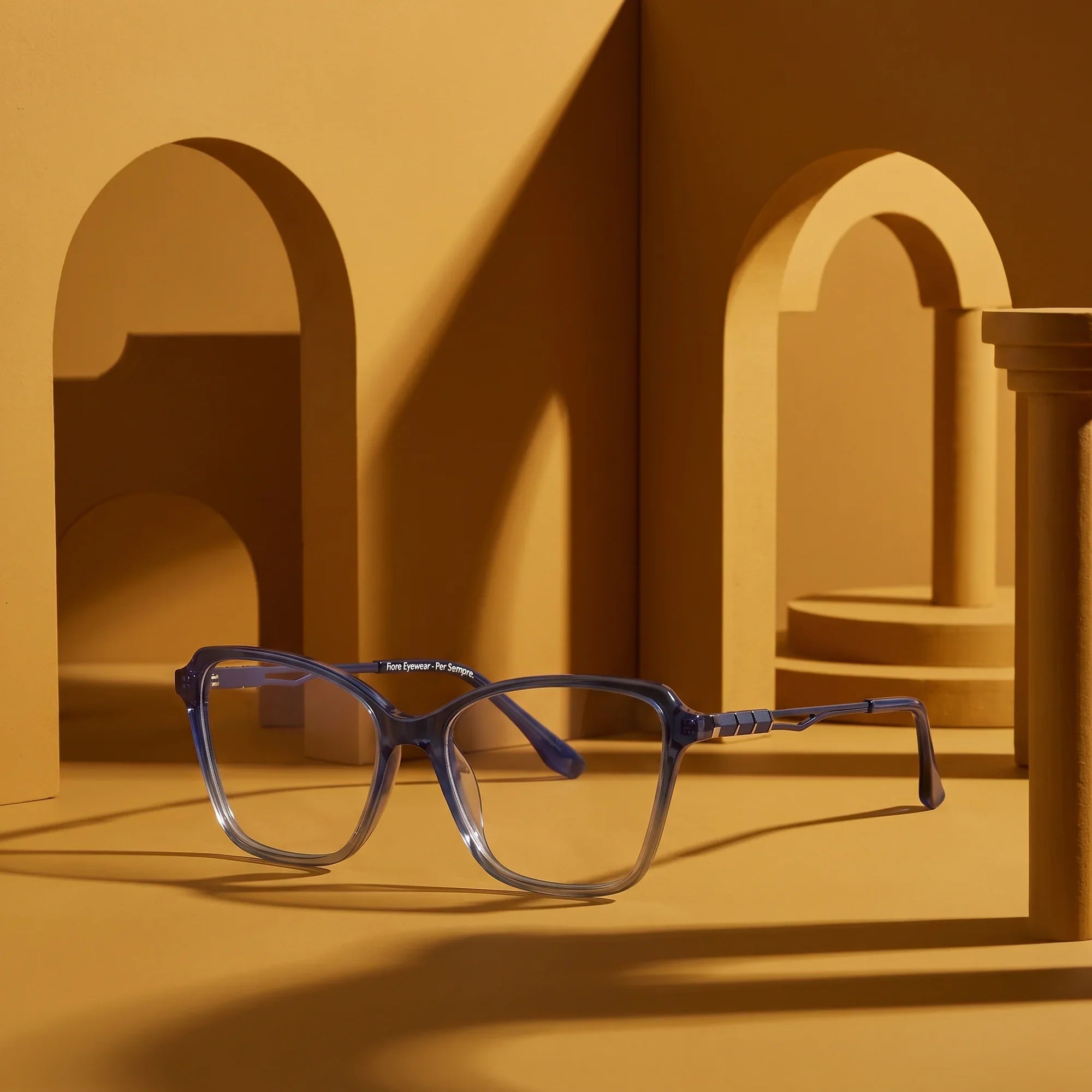 Anteojos Fiore 1006 en azul – displayed on a mustard ochre podium among arches
