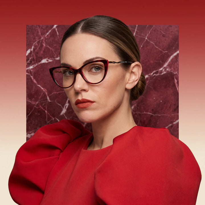 Anteojos Fiore 1010 en rojo, en modelo – retrato de campaña, fondo de mármol rojo y vestido escarlata drapeado