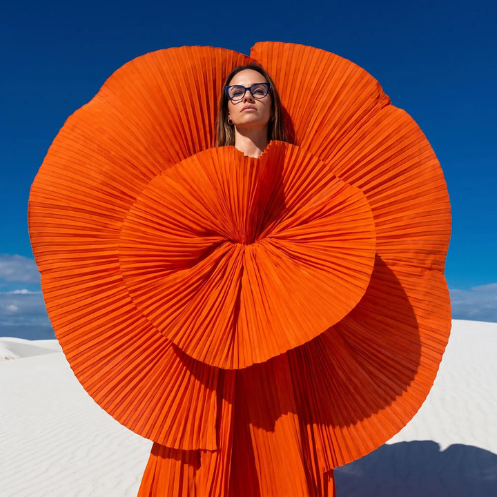 Anteojos Fiore 1010 en azul, en modelo – retrato de campaña, abanico plisado naranja escultórico sobre dunas blancas