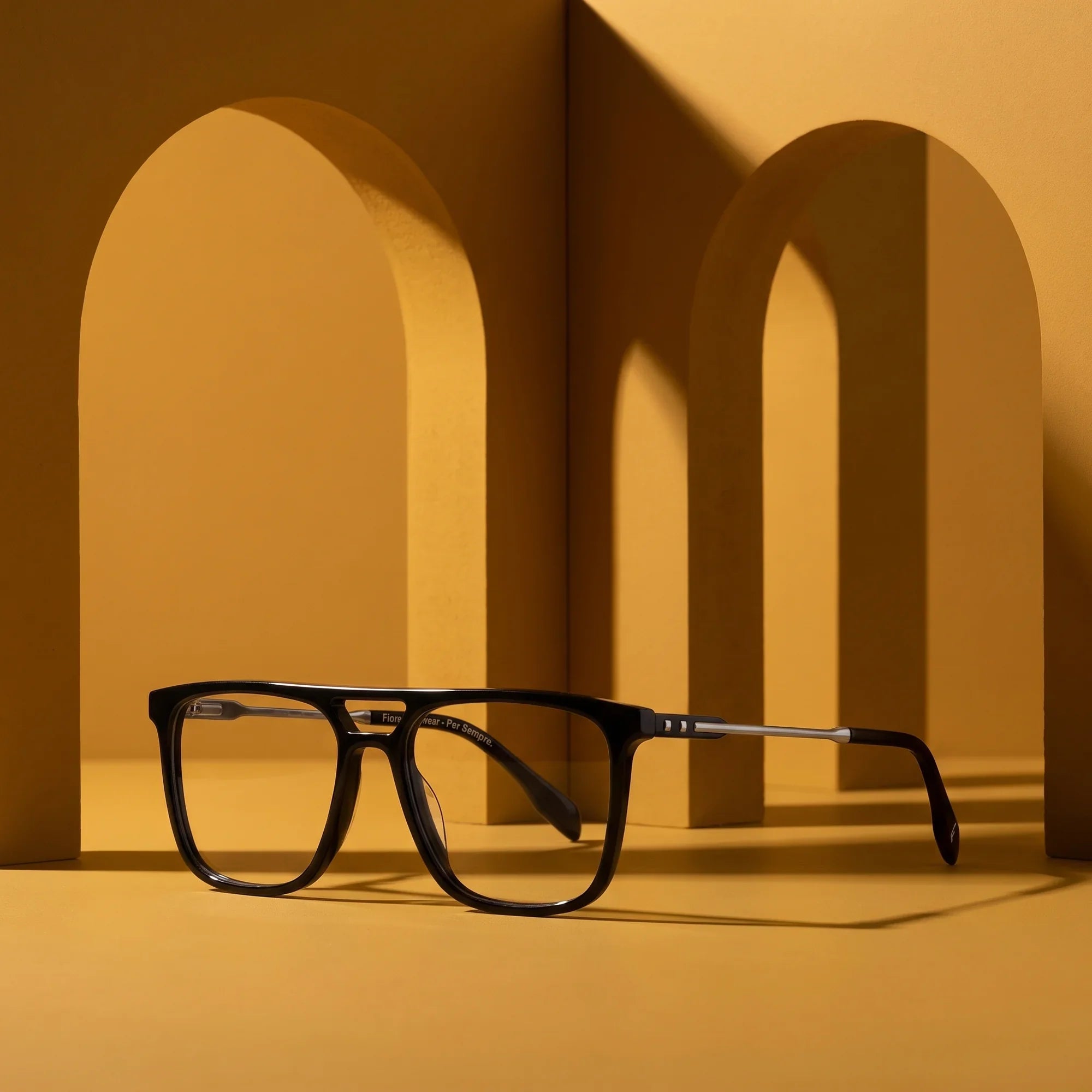Anteojos Fiore 10177 en negro – displayed on a mustard ochre podium among arches