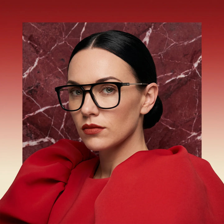 Anteojos Fiore 10177 en negro, en modelo – retrato de campaña, fondo de mármol rojo y vestido escarlata drapeado