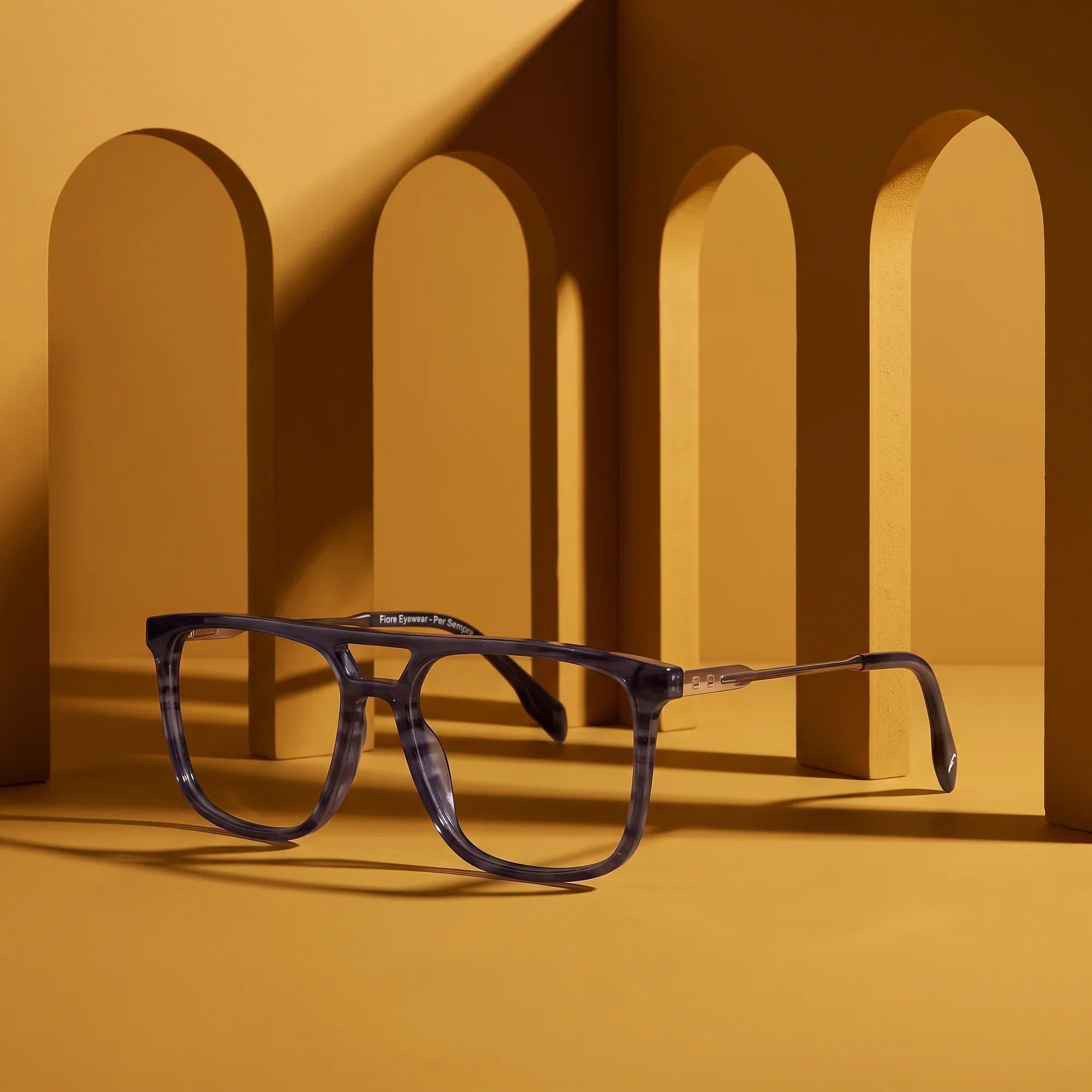 Anteojos Fiore 10177 en gris – displayed on a mustard ochre podium among arches