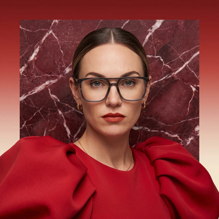 Anteojos Fiore 10177 en gris, en modelo – retrato de campaña, fondo de mármol rojo y vestido escarlata drapeado