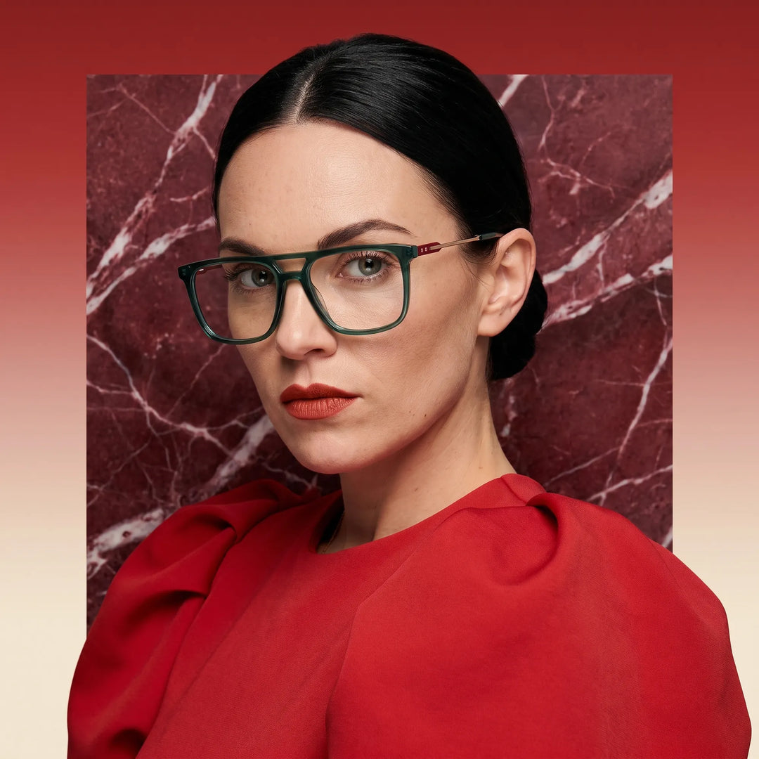 Anteojos Fiore 10177 en verde, en modelo – retrato de campaña, fondo de mármol rojo y vestido escarlata drapeado