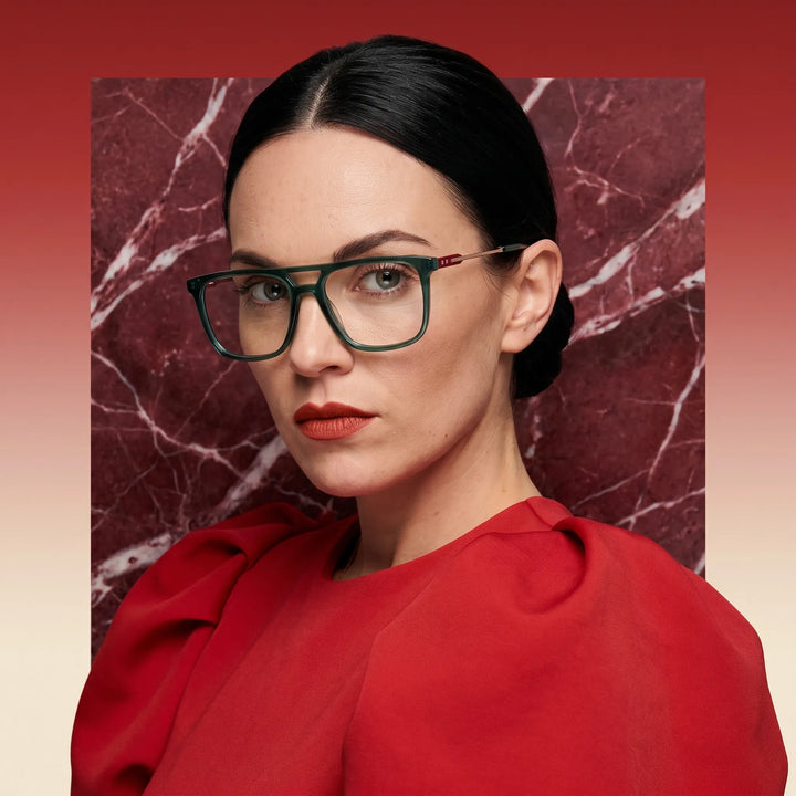 Anteojos Fiore 10177 en verde, en modelo – retrato de campaña, fondo de mármol rojo y vestido escarlata drapeado