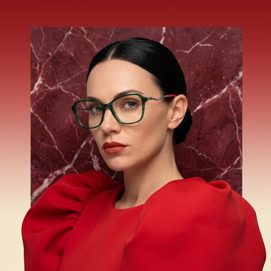 Anteojos Fiore 1030 en verde, en modelo – retrato de campaña, fondo de mármol rojo y vestido escarlata drapeado