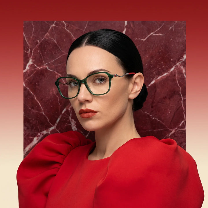 Anteojos Fiore 1030 en verde, en modelo – retrato de campaña, fondo de mármol rojo y vestido escarlata drapeado