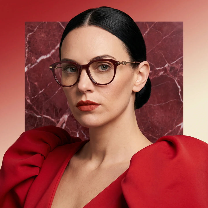 Anteojos Fiore 5409 en brown red, en modelo – retrato de campaña, fondo de mármol rojo y vestido escarlata drapeado