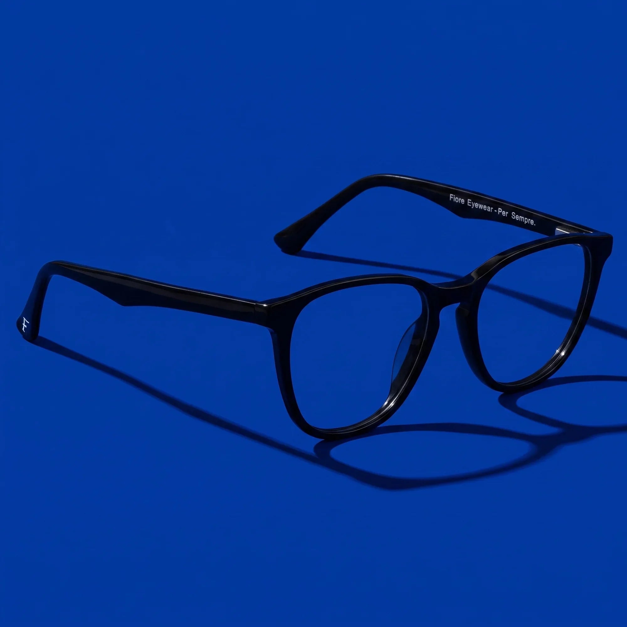 Anteojos Fiore 7046 en negro – hero still life against a deep cobalt backdrop