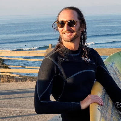 un modelo hombre en la playa con traje de surf y sosteniendo una tabla de surf usando lentes hexagonales color carey con lentes polarizadas. El hombre está sonriendo y de fondo se ve el mar de la playa pichilemu